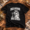 Schwarzes Pferdebua-Herrenshirt mit Pferd, Sonnenbrille, Mikrofon und „Mei Bühne is mei Stall“ auf einem Holztisch