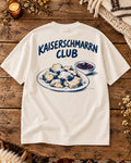 Weißes Oversize-T-Shirt Kaiserschmarrn Club auf einem Altholztisch – Rückenmotiv