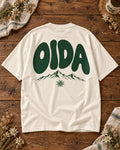 Weißes Oversize-T-Shirt OIDA mit grünem Bergmotiv und Edelweiß – Rückseite auf Altholztisch