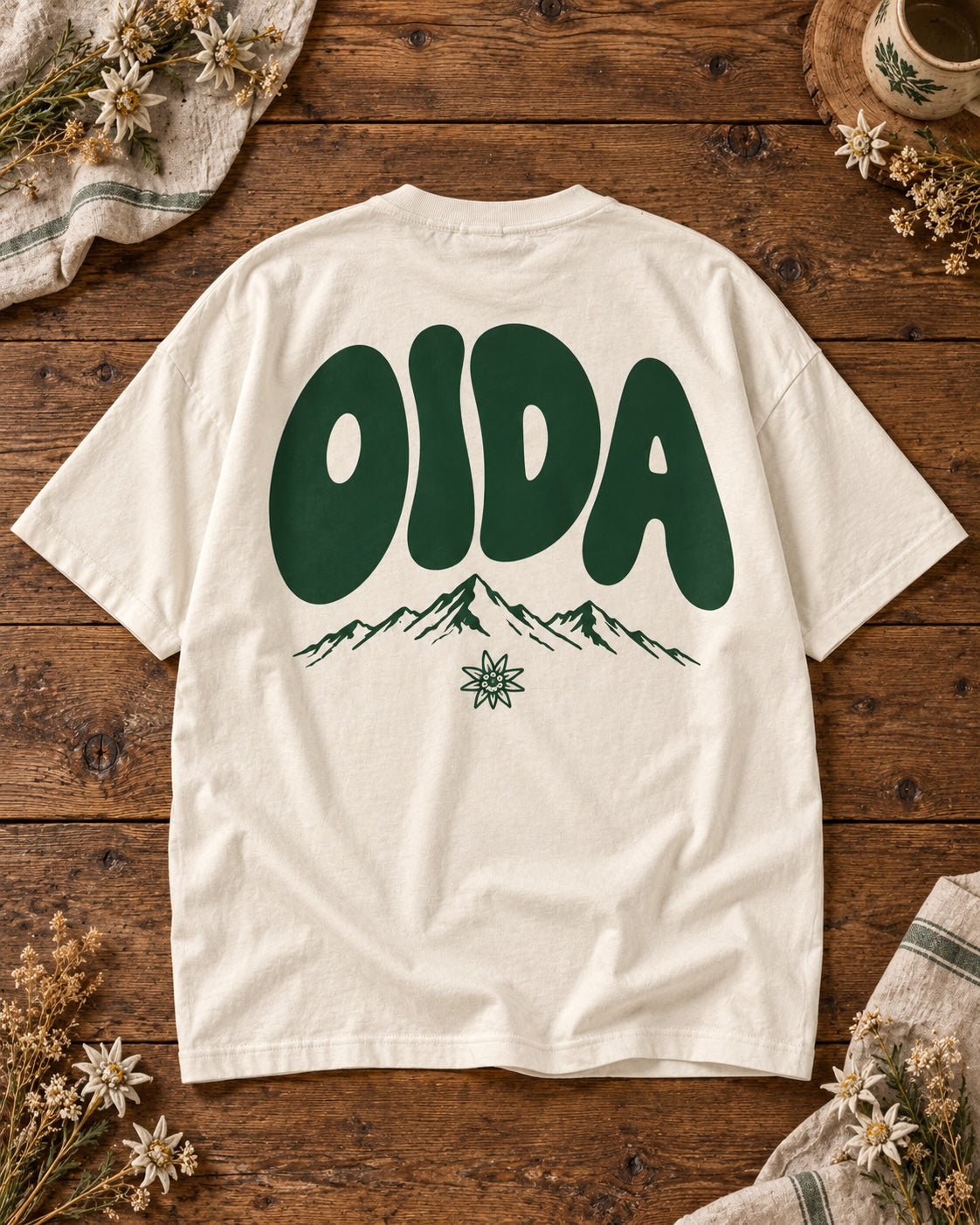 Weißes Oversize-T-Shirt OIDA mit grünem Bergmotiv und Edelweiß – Rückseite auf Altholztisch