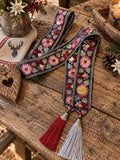 SCHMIGI Taillengürtel Navy mit buntem Blumenmuster und roten und hellgrauen Quasten auf einem Altholztisch
