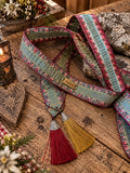SCHMIGI Taillengürtel Türkis mit Blumenbordüren und roten und goldgelben Quasten auf einem Altholztisch